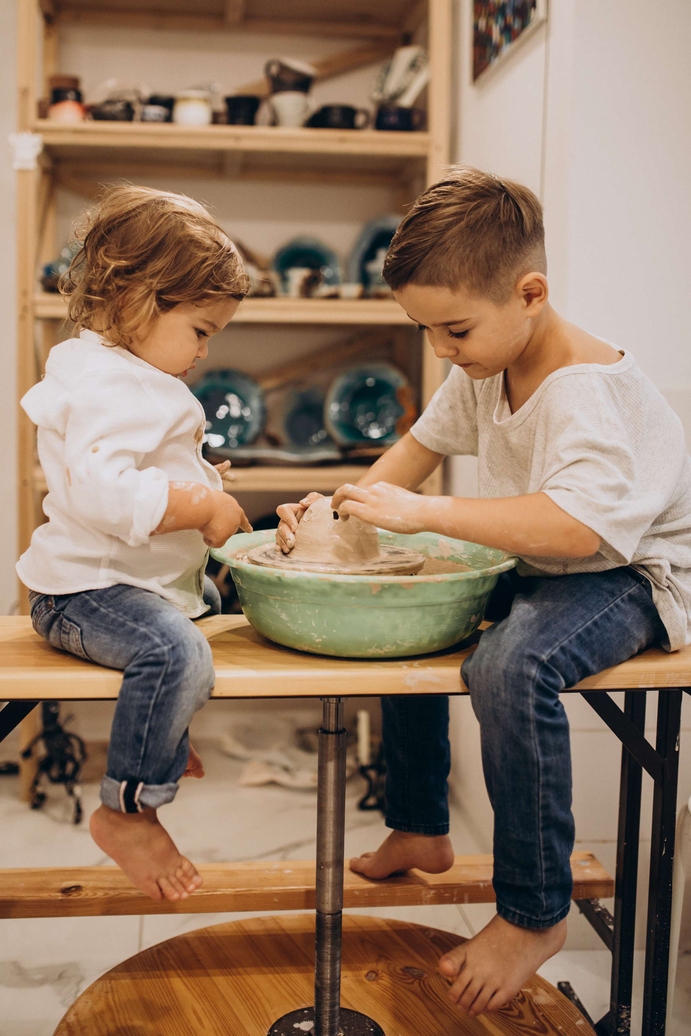 Two young brothers work together shaping clay at a small pottery wheel during a kids pottery class.