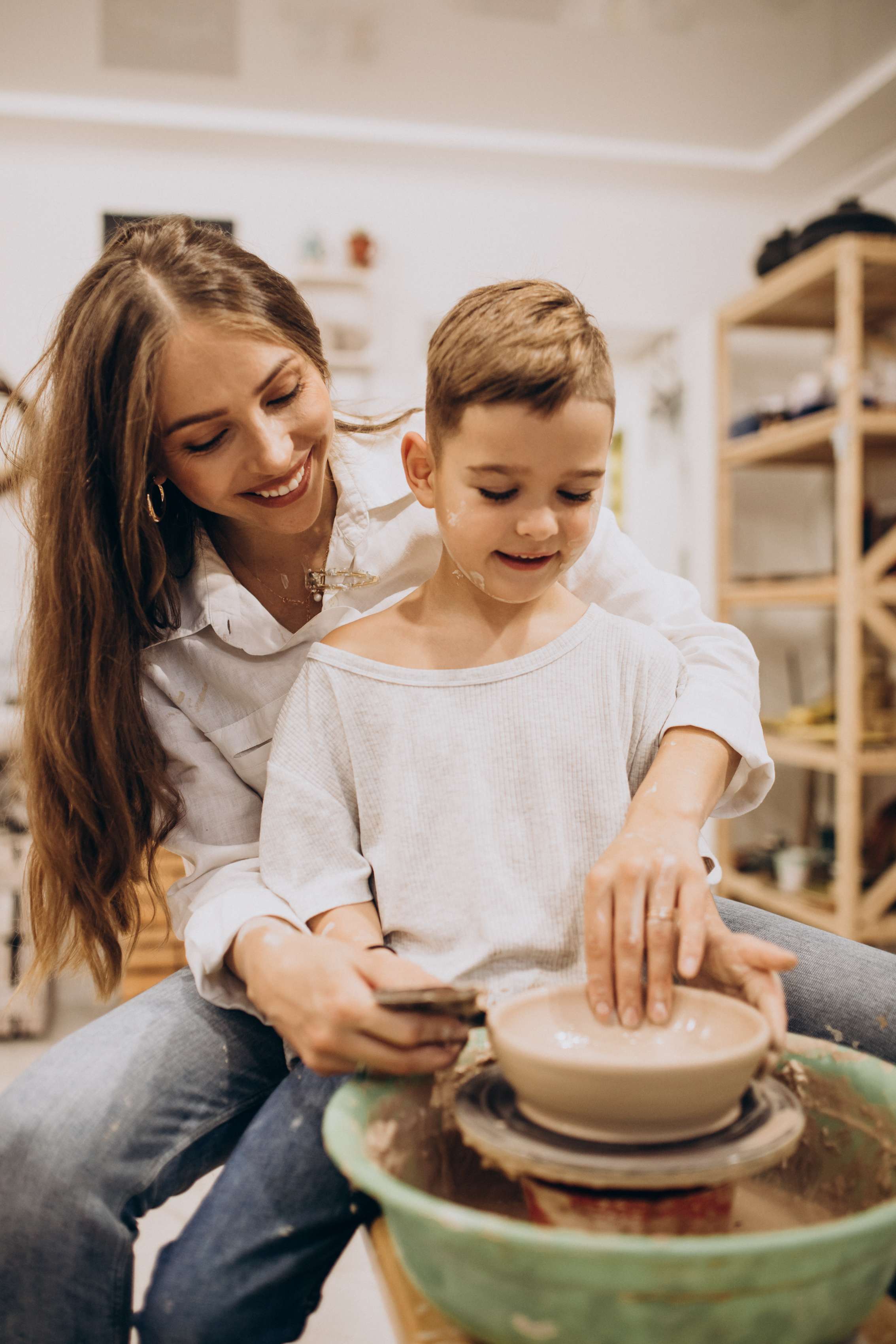 A smiling mother guides her young son's hands on a pottery wheel during a family pottery class.