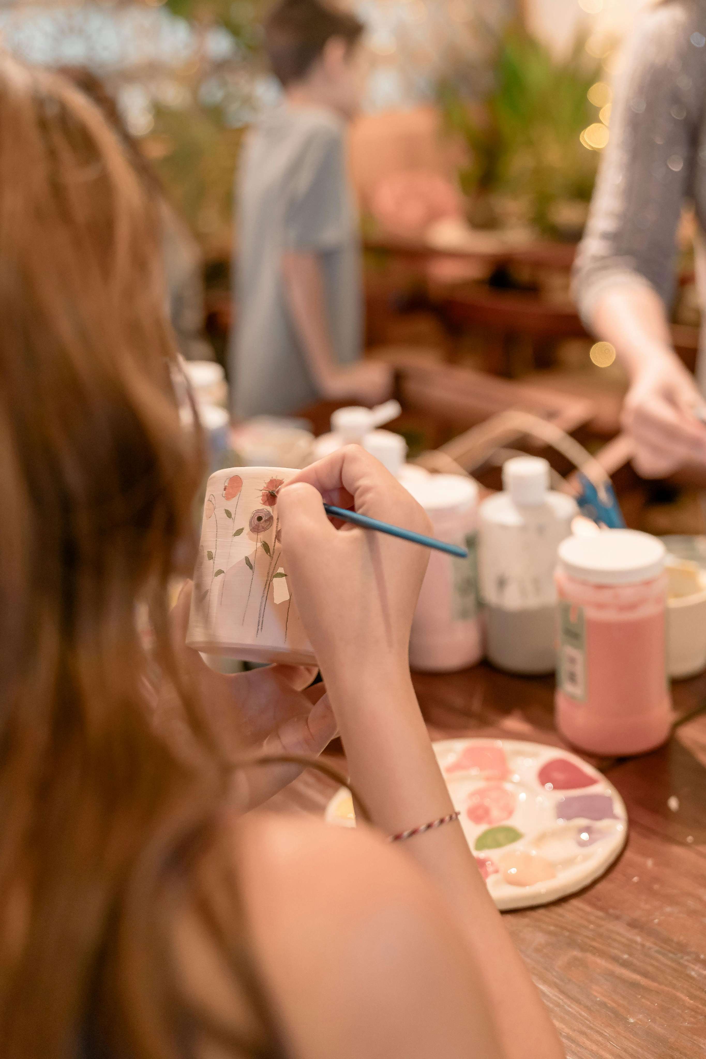 A student using a fine brush to paint floral designs on a ceramic mug during a pottery class.