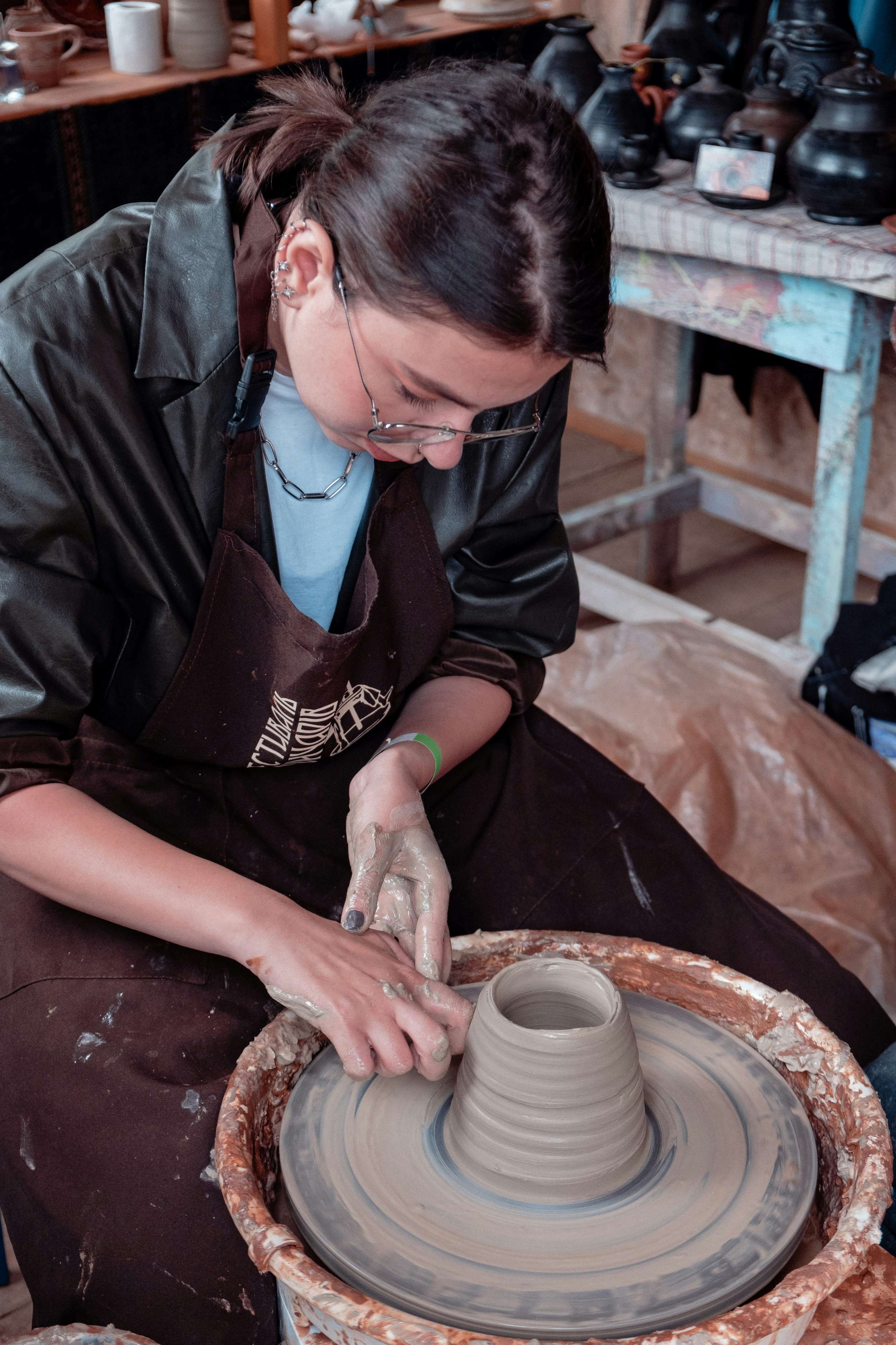 A woman in a brown apron concentrates as she throws clay on a pottery wheel in a ceramics studio.
